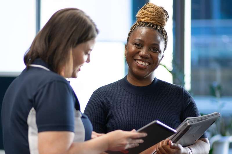 Two students standing in the library holding laptops whilst one smiles at the camera