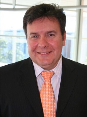 John Kapeleris smiling, wearing a black suit coat, light orange and blue business shirt and bright orange tie