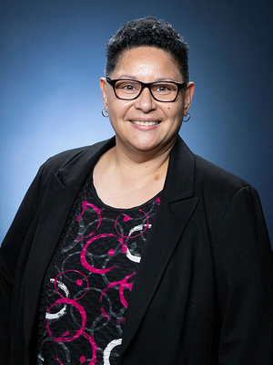 Leilani Bin-Juda smiling with black reading glasses, wearing a black suit coat with pink and white circles top