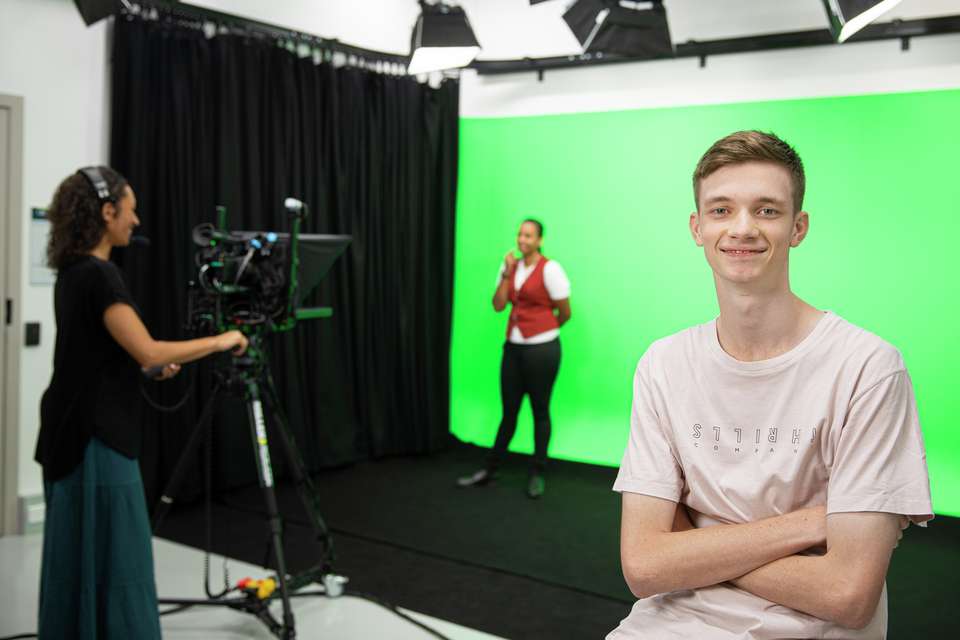 A digital media student sitting and smiling in a studio while two students work with a green screen in the background