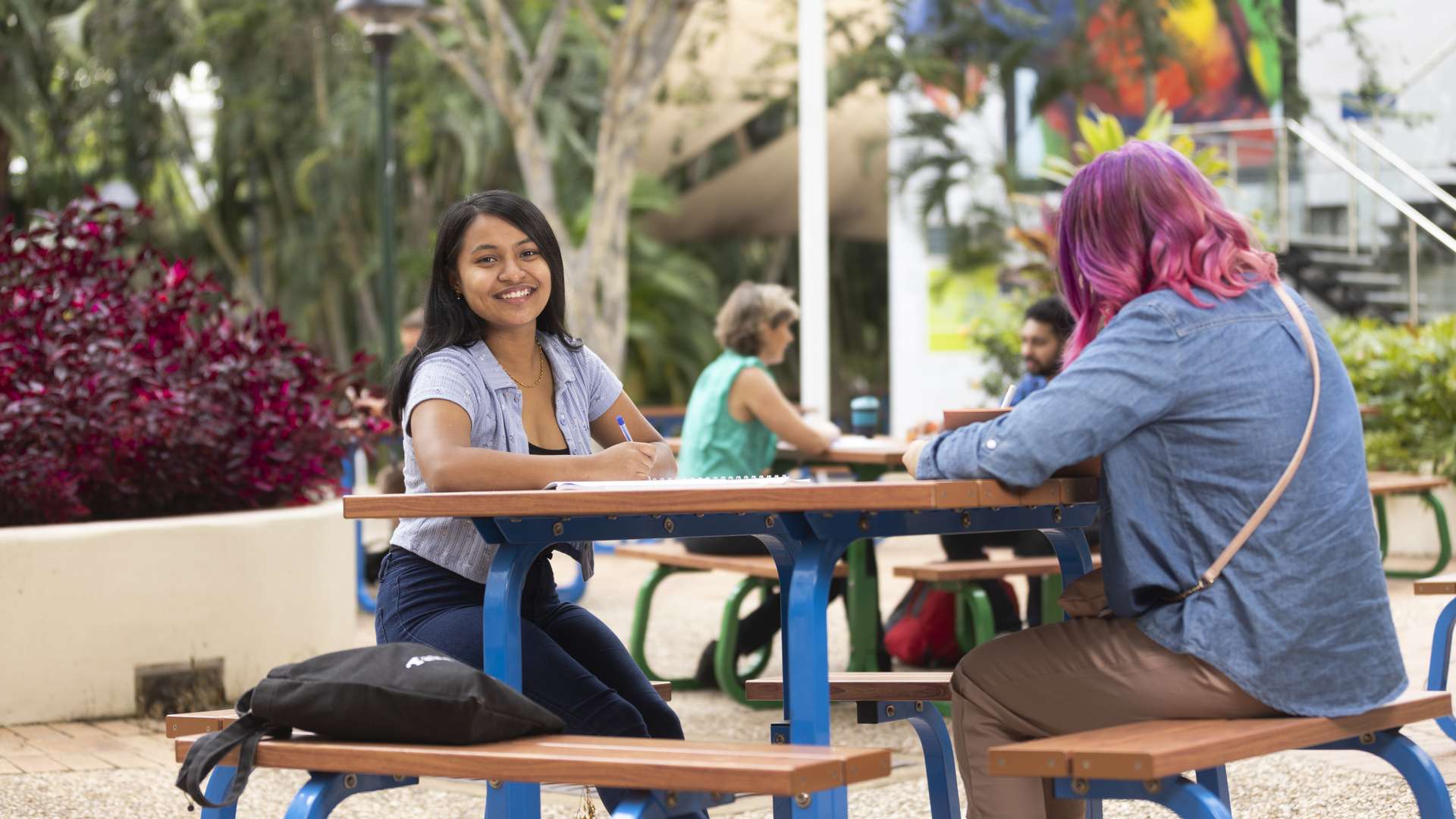 Smiling student studying at outdoor table with another student during the day.