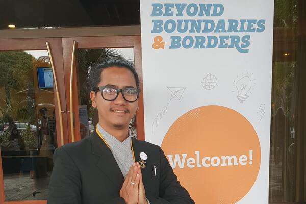 Bikesh Lal Shresthas standing in front of a Welcome sign greeting with a namaste gesture