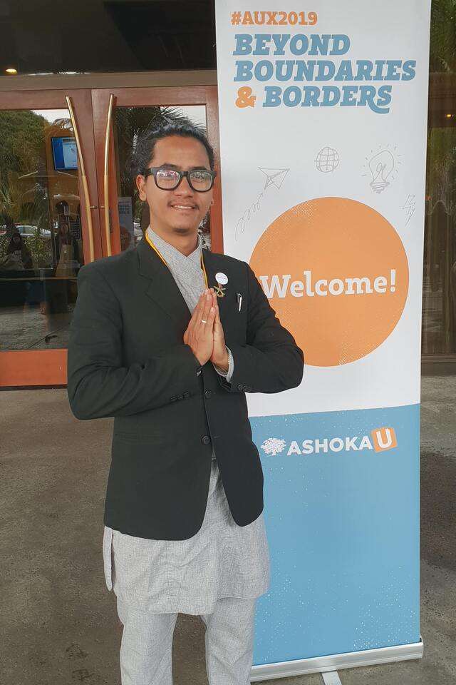 Bikesh Lal Shresthas standing in front of a Welcome sign greeting with a namaste gesture
