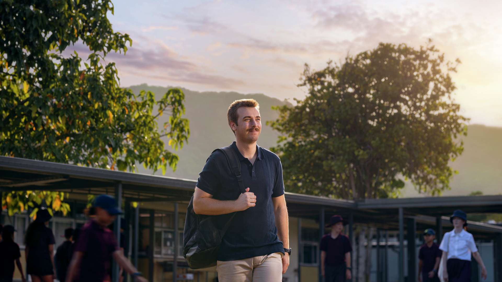 Student teacher walks outdoors on a sunny day, carrying a bag over his shoulder. Trees and school buildings are in the background, with other people walking nearby.