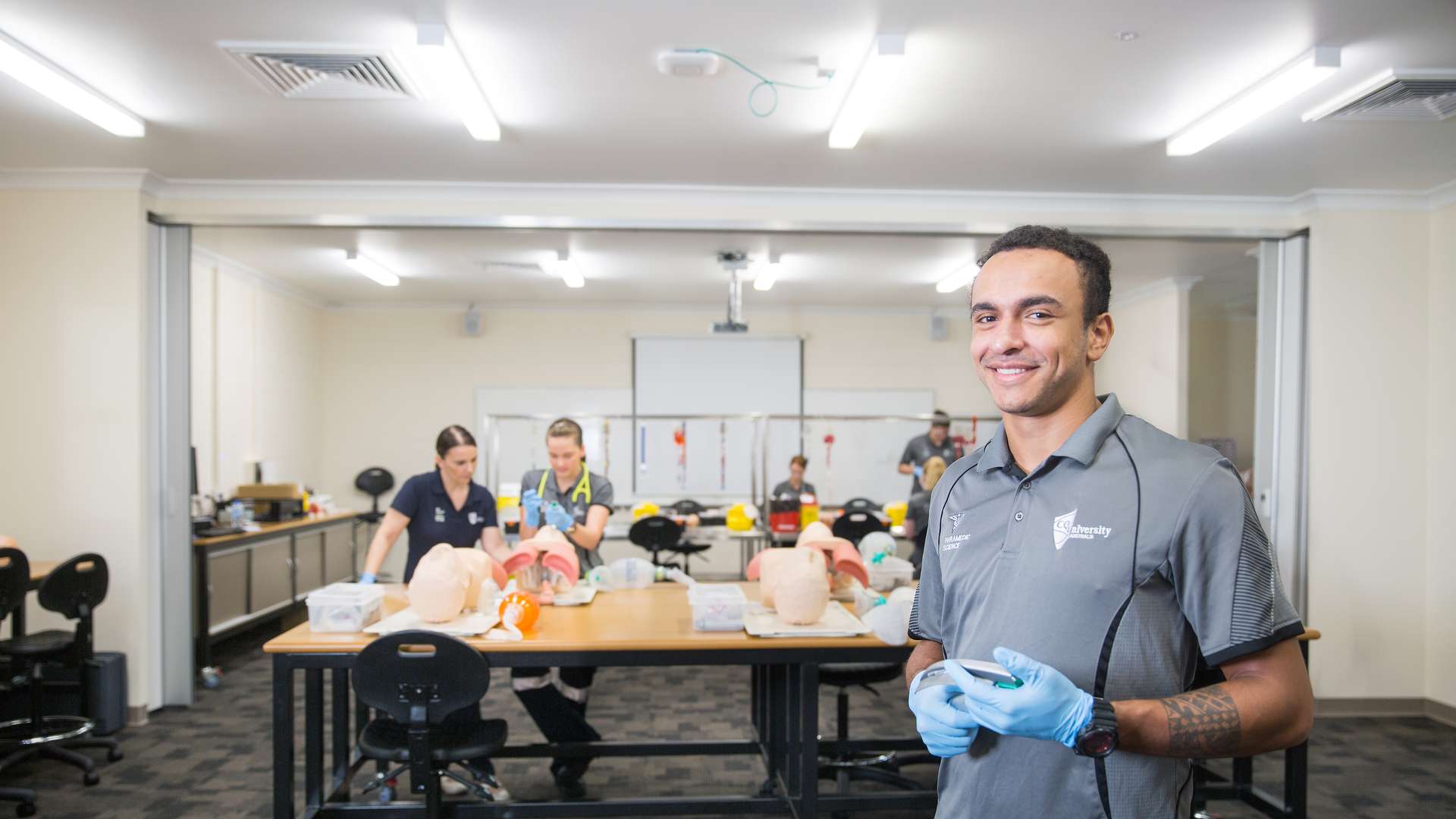 Paramedic science student smiles at camera while students and lecturer work in the background.
