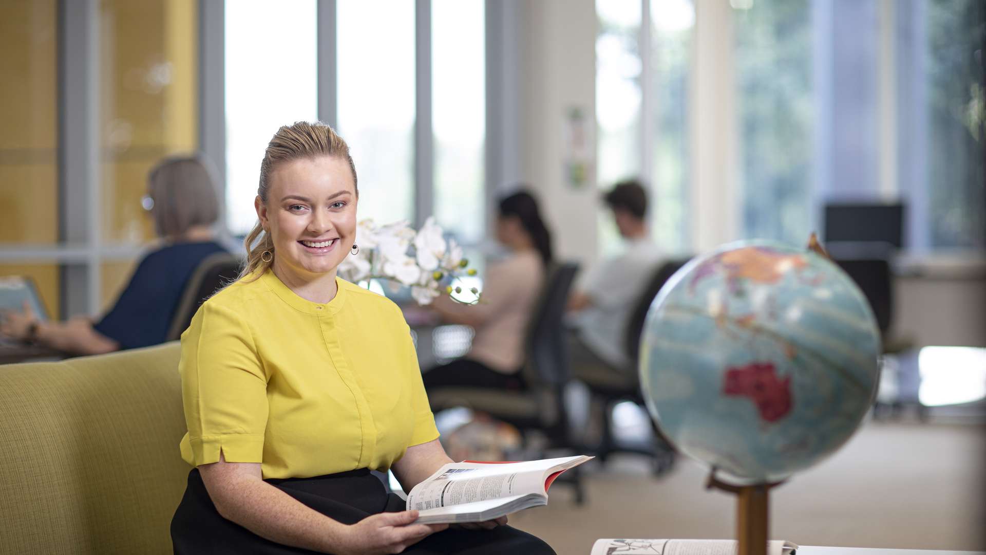 A student sitting on a lounge holding a textbook. A globe of the Earth is in the foreground, out of focus and more students can be seen in the background studying.