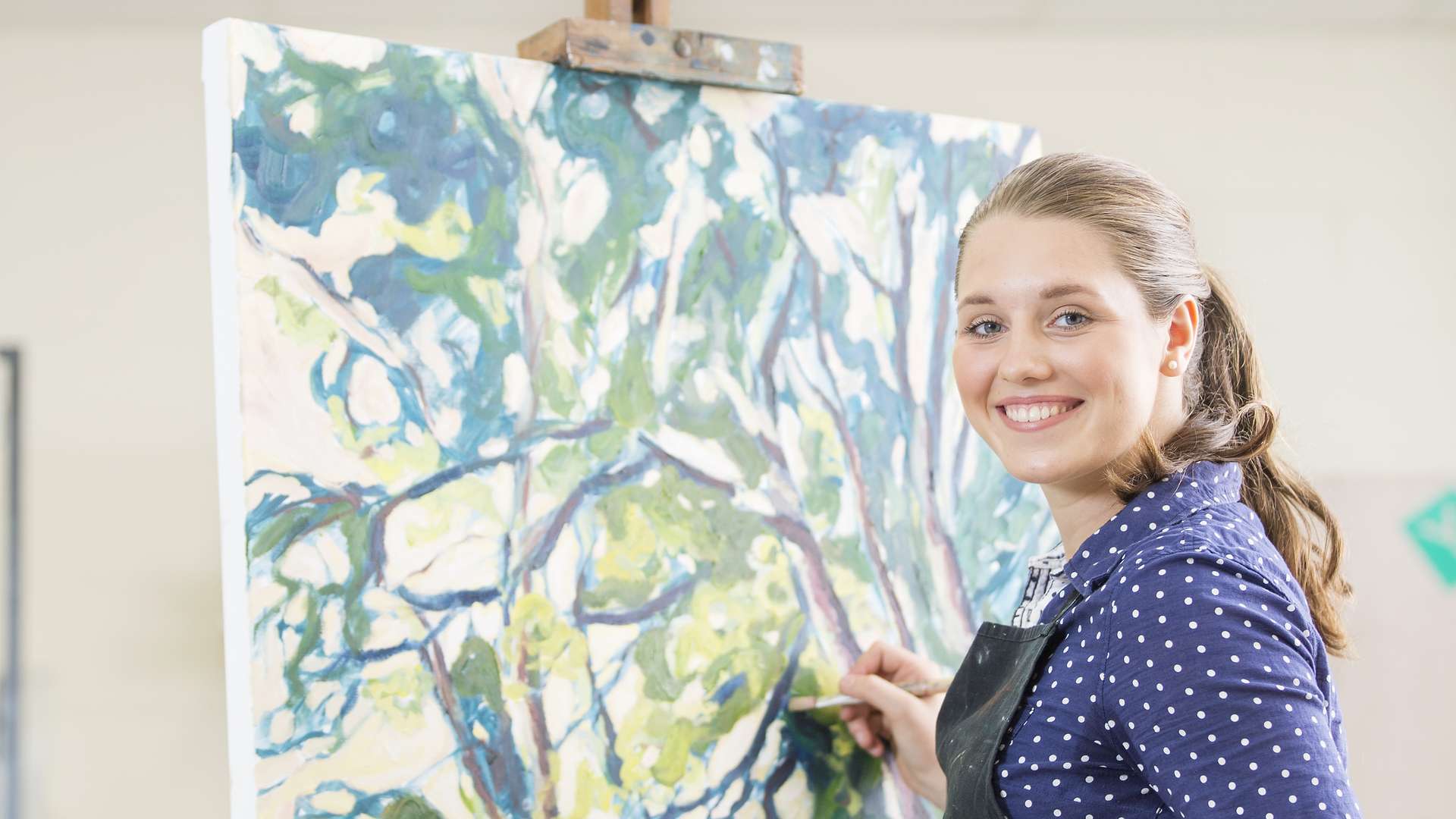 Art student smiling as she paints her artwork in the background.