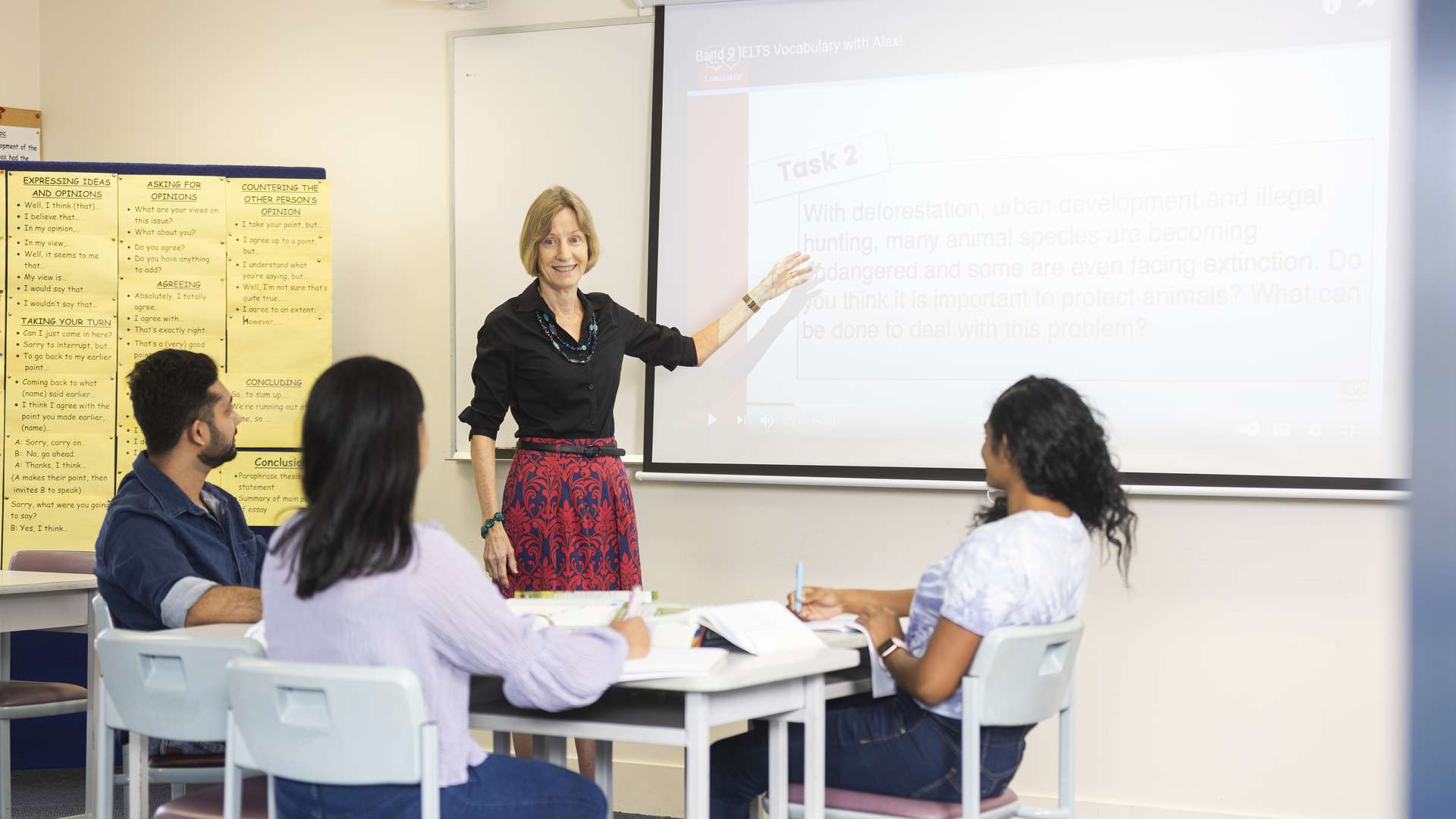 Three CQU English students sitting at a desk with workbooks and textbooks whilst a tutor points towards a slideshow..