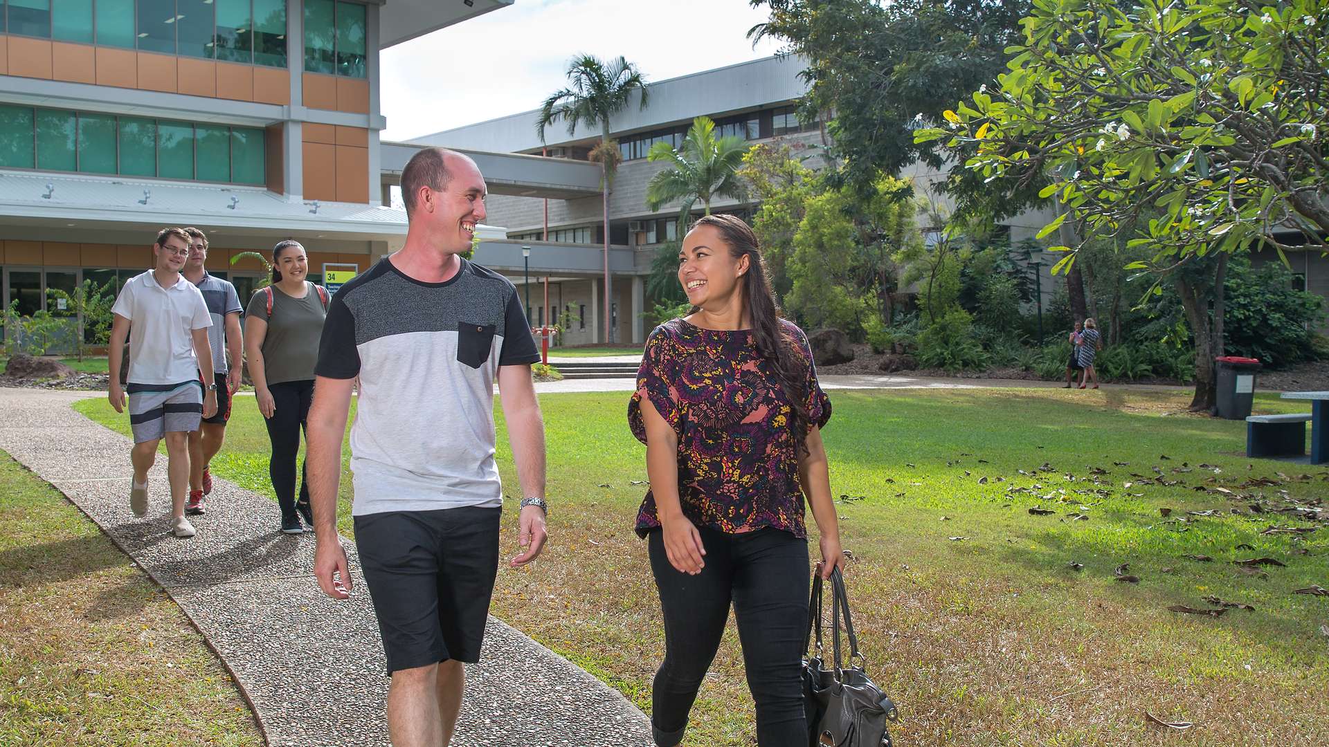 Two smiling students walking on path, one holding a bag with other students walking in background.