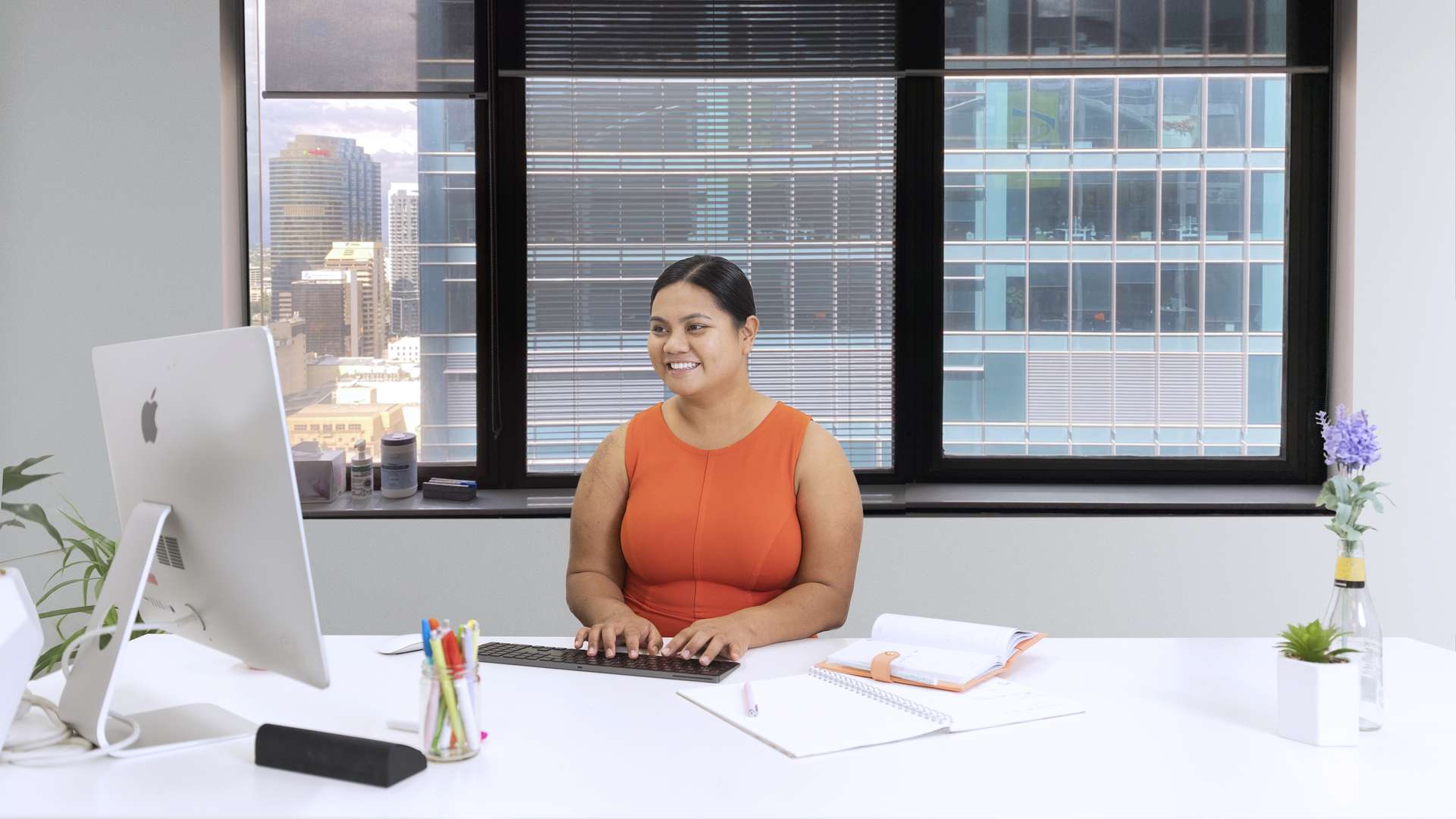 Student working in office smiling towards computer with window view of building outside