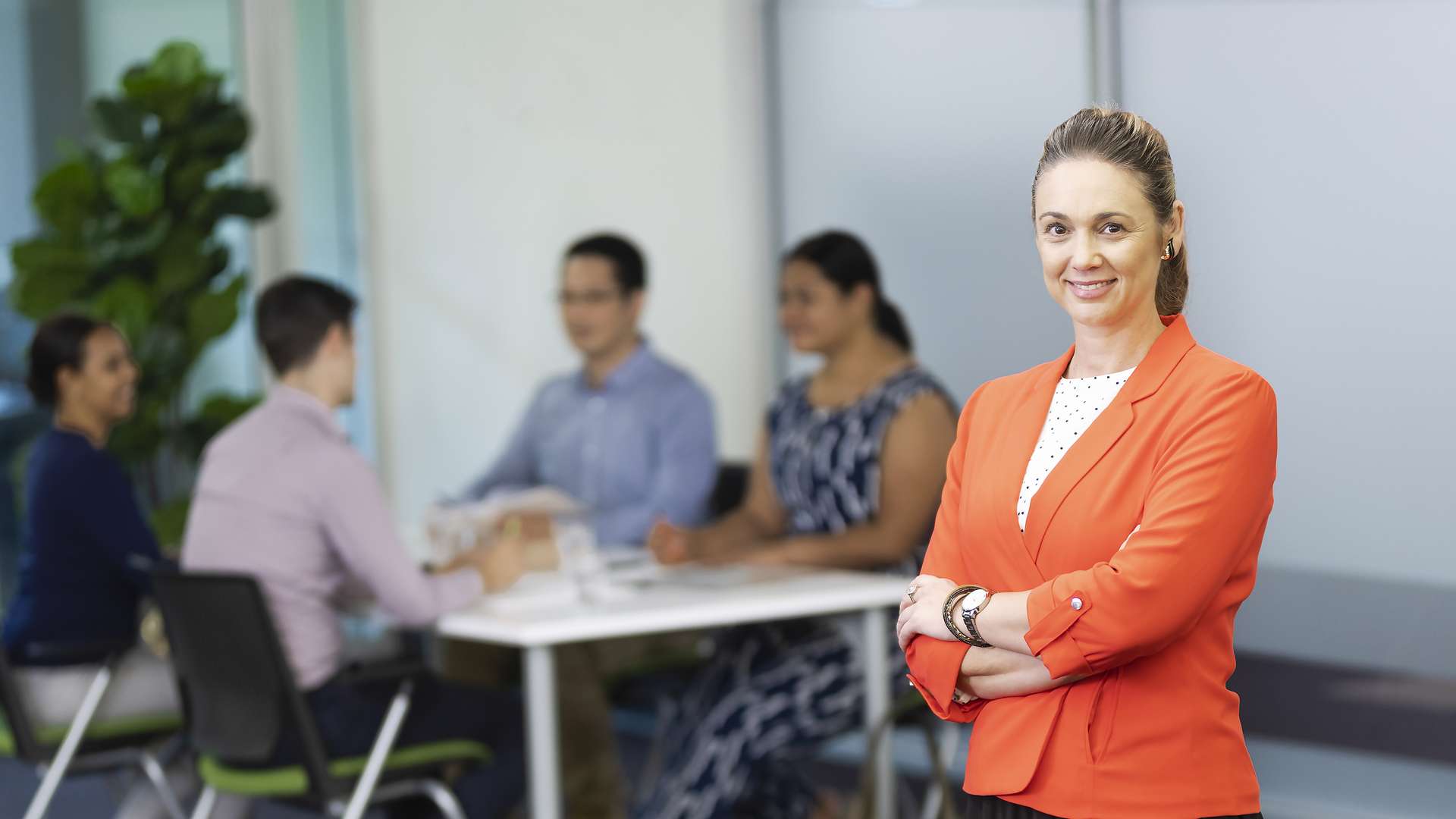 A woman, dressed professionally, standing in the foreground with her arms crossed, with a group people at a table behind her