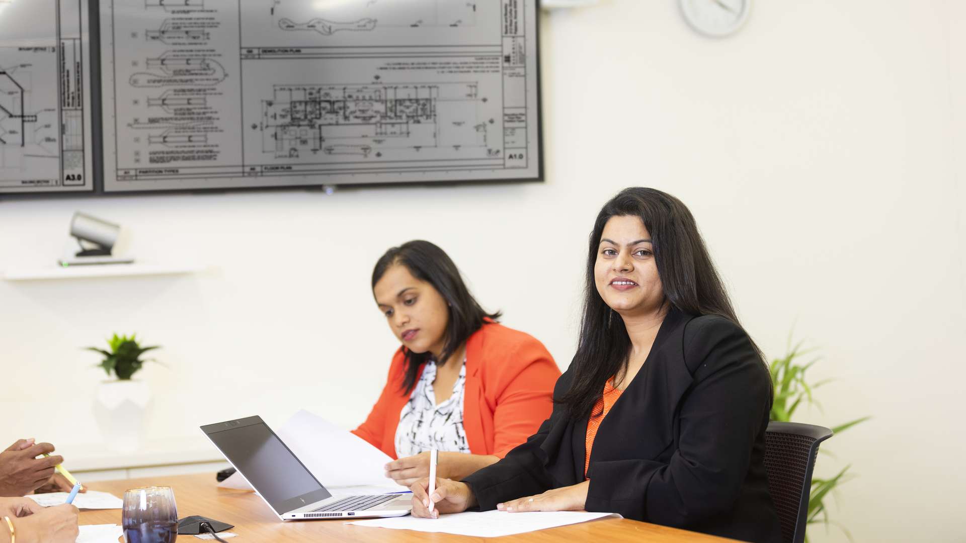 Two construction students working together at a desk. Construction plans are displayed on two monitors in the background.