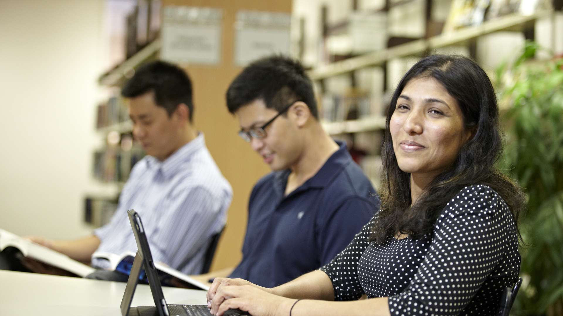 Three students studying in the library, two reading books and the third using an iPad.