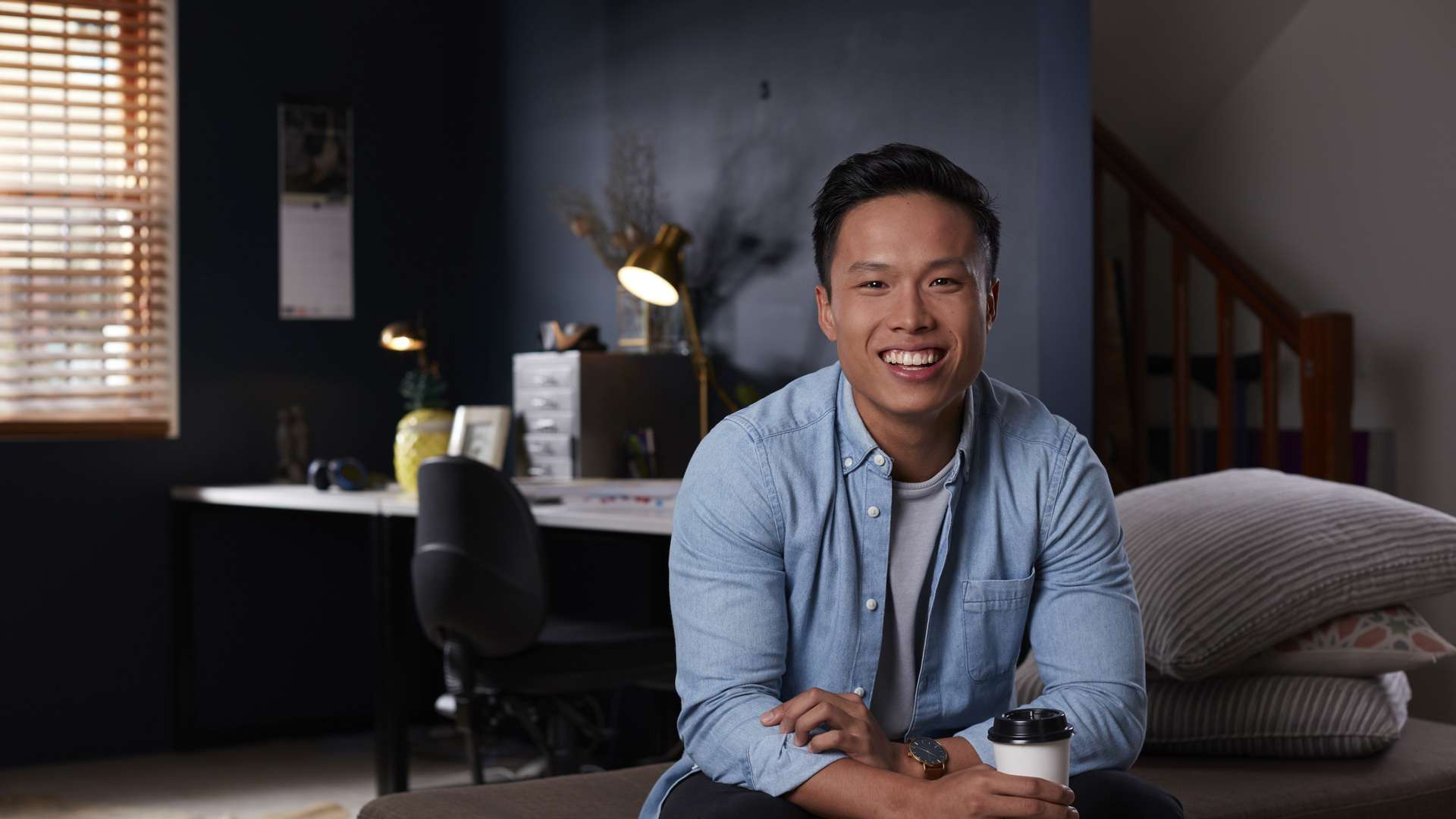 An online student sitting on a couch smiling and holding a takeaway coffee