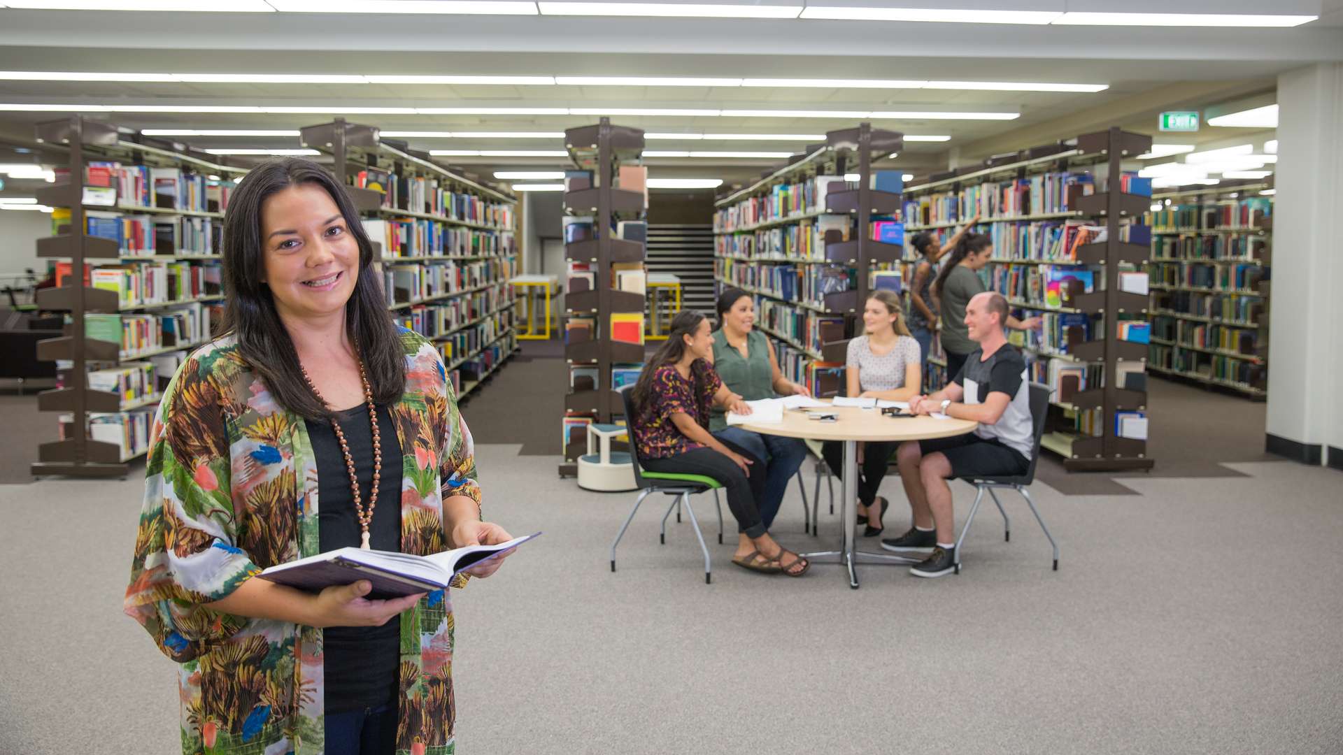 Students seated around a library table as creative writer stands smiling with a book.