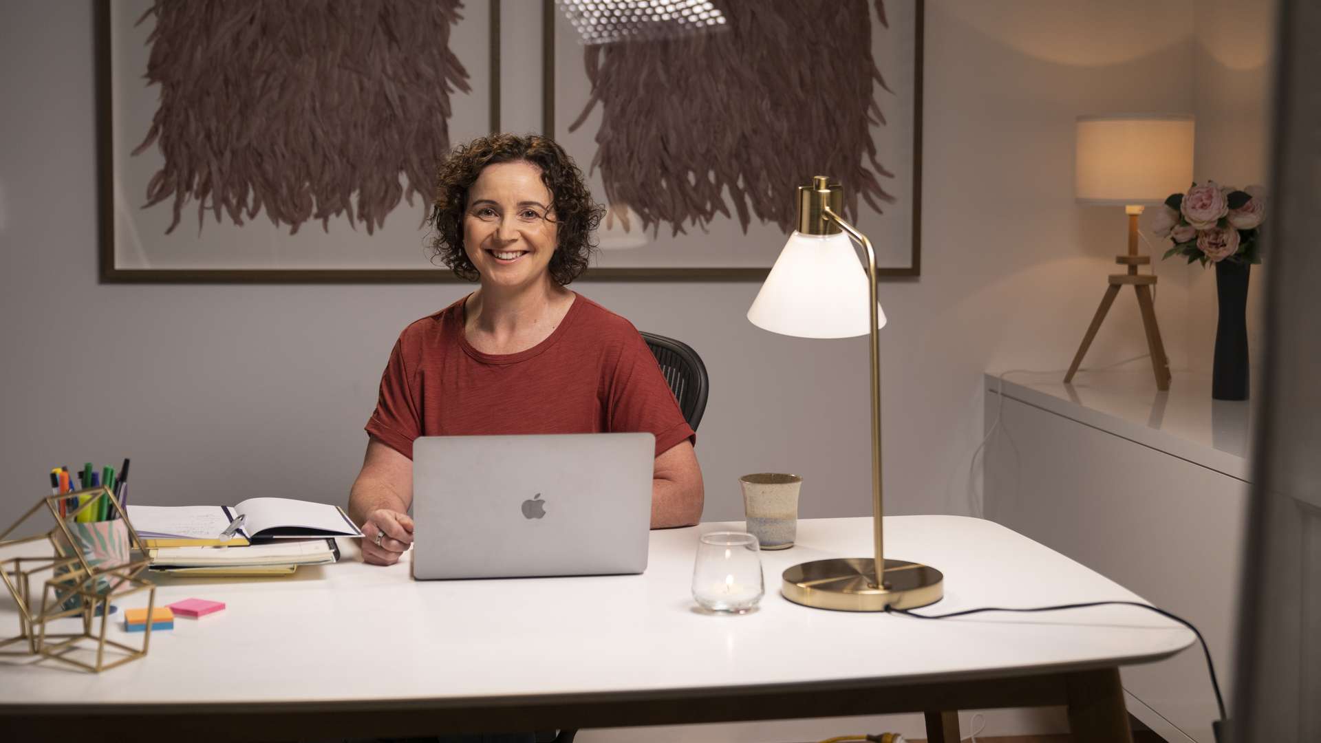 A lady sitting at a desk with a workbook and laptop. Two paintings are mounted behind the person on the wall.