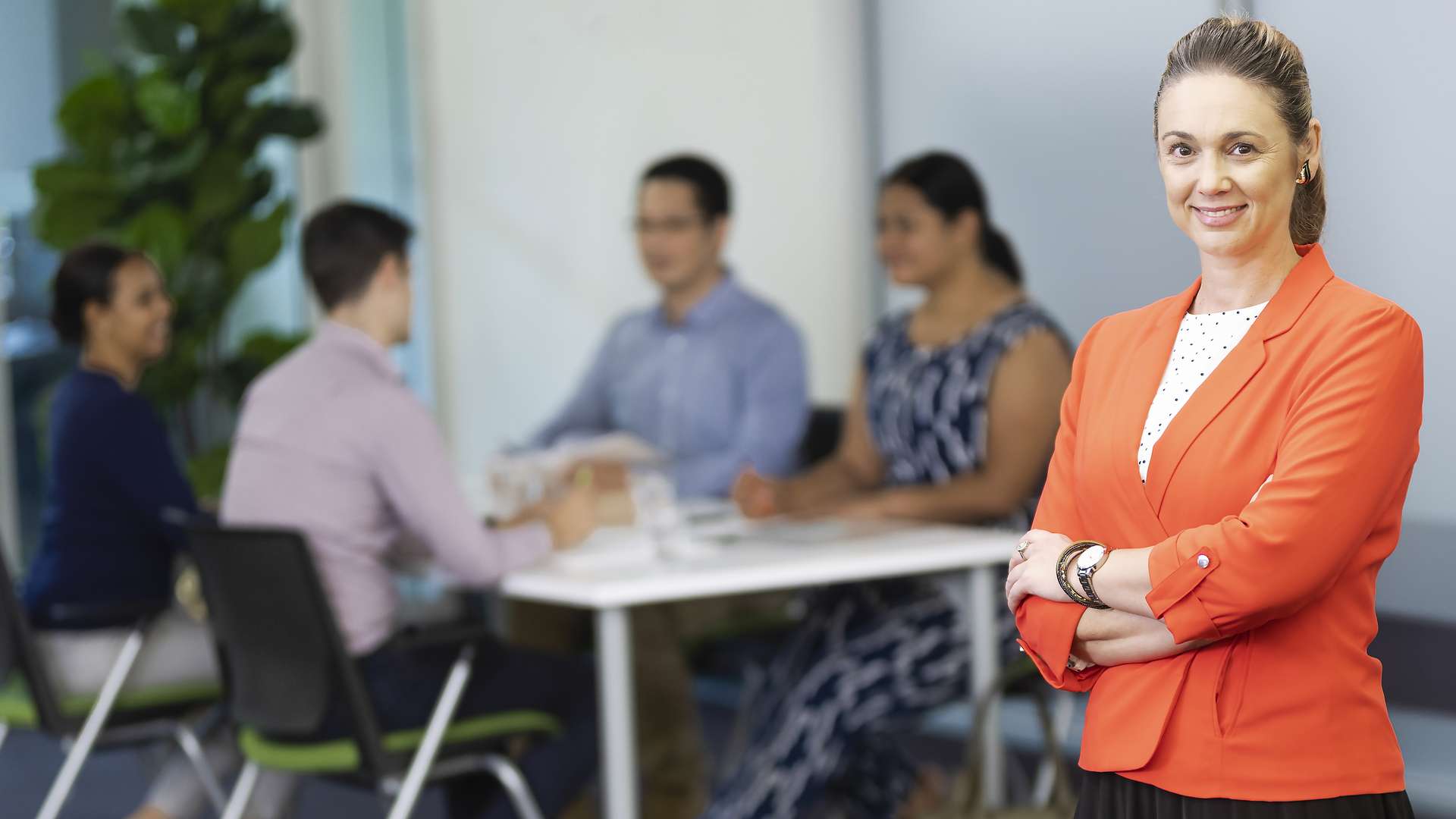 A woman, dressed professionally, standing in the foreground with her arms crossed, with a group people at a table behind her