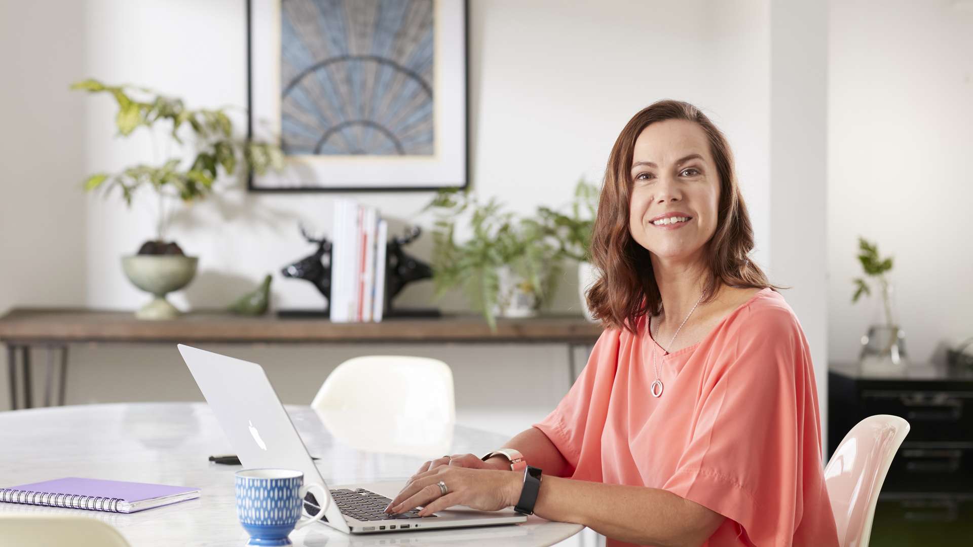 A student sitting at a table at home using their laptop whilst having a cup of tea.