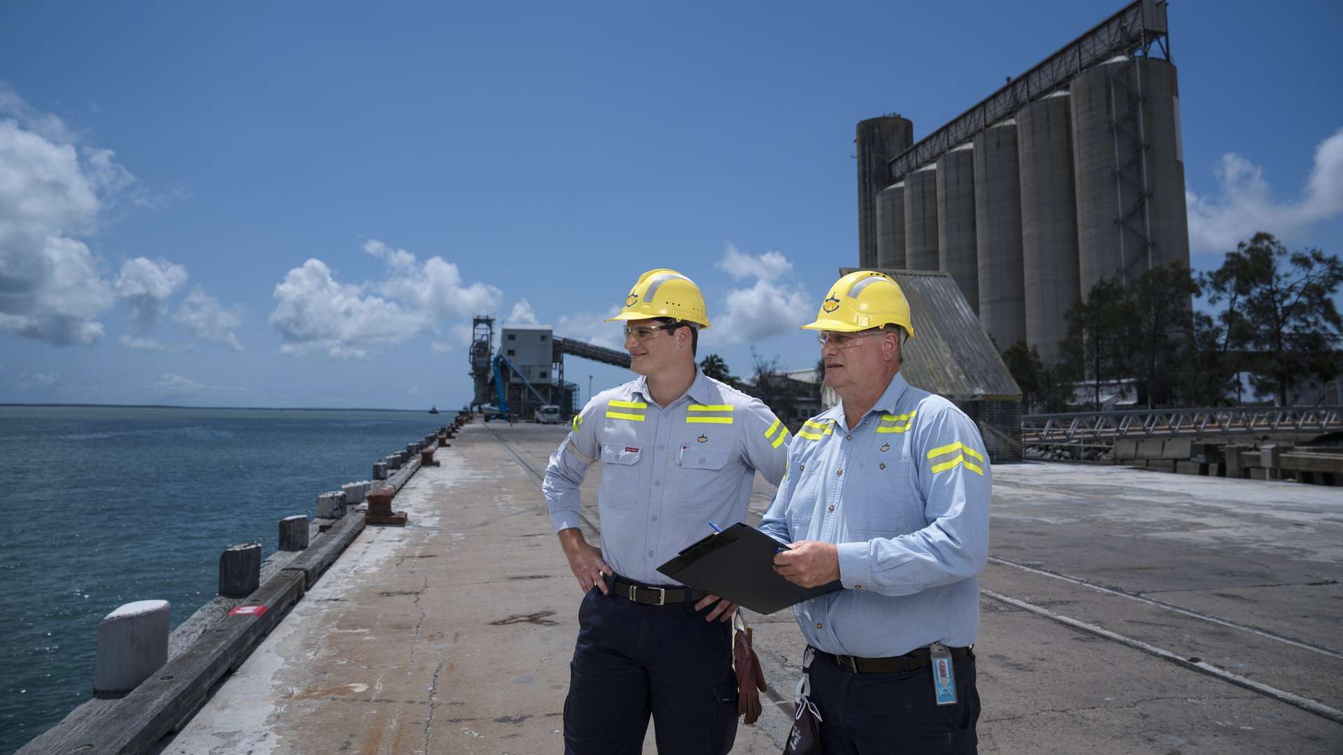 Two men in hard hats standing on the edge of an industrial shipping port, the ocean and a silo are in the background.