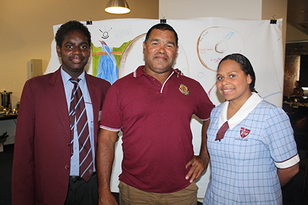 Two Indigenous males and one Indigenous female standing in front of an Indigenous artwork