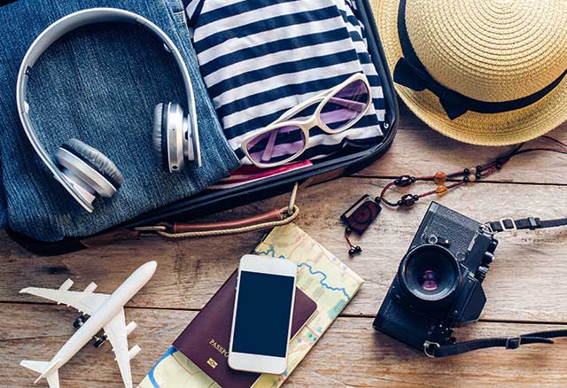 Flat lay photo of a camera, phone, passport, map, bead necklace toy airplane, straw hat, blue jeans, striped shirt, headphones and sunglasses