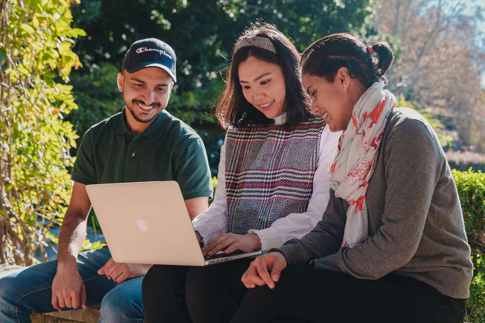 Three students sitting outside, the outer two students focusing on the centre student using their laptop