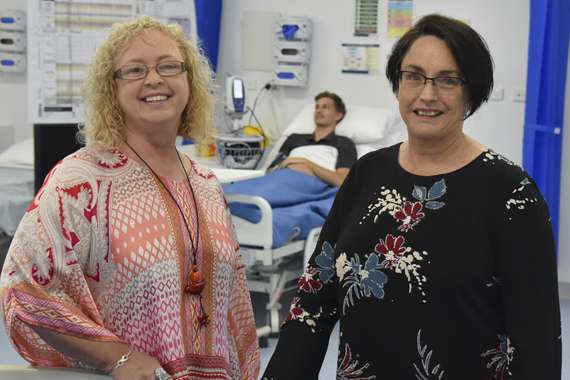 Two women smiling in a hospital in front of a patient in a hospital bed