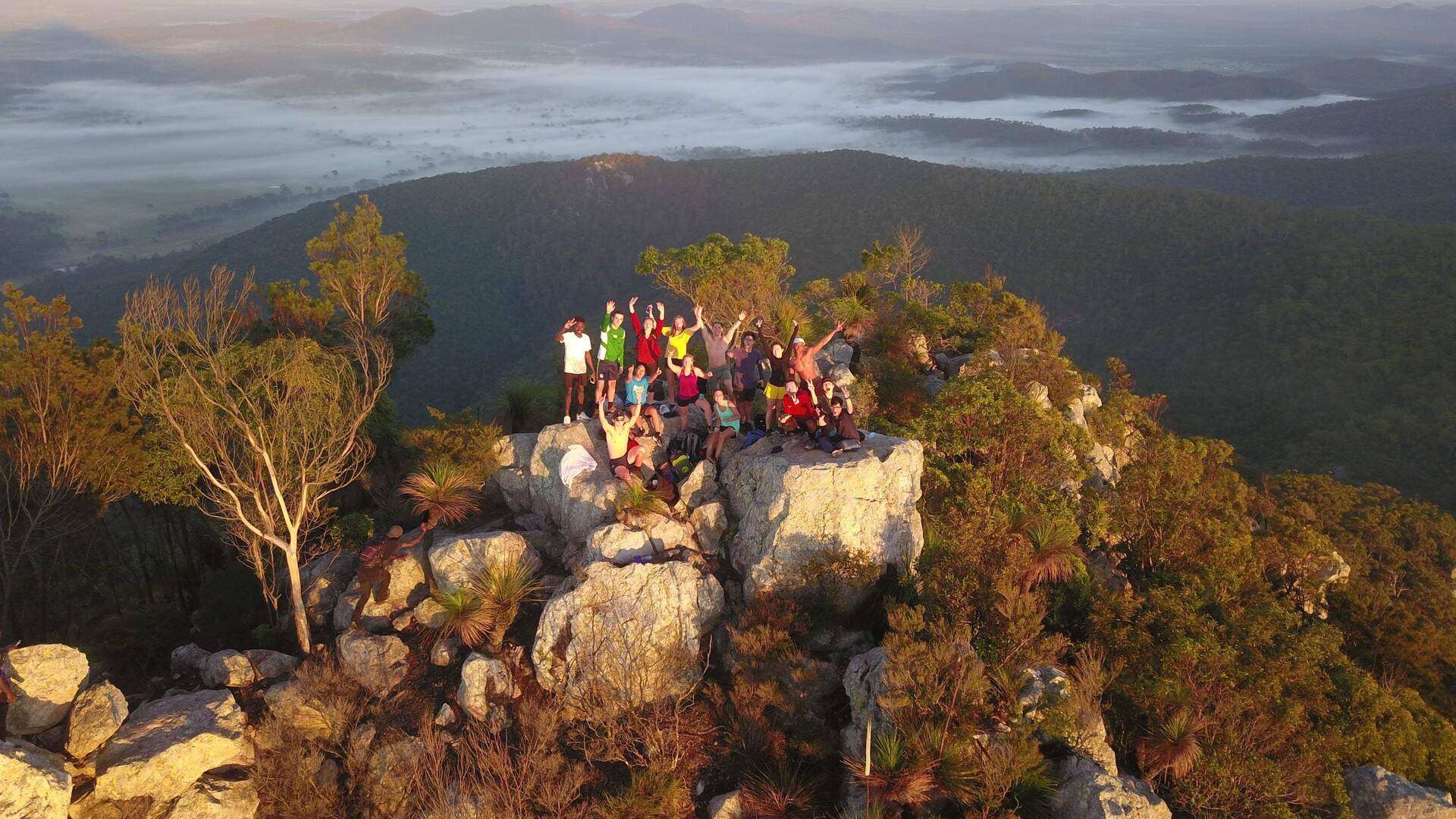 An aerial view of students standing on the summit of a mountain, their hands in the air