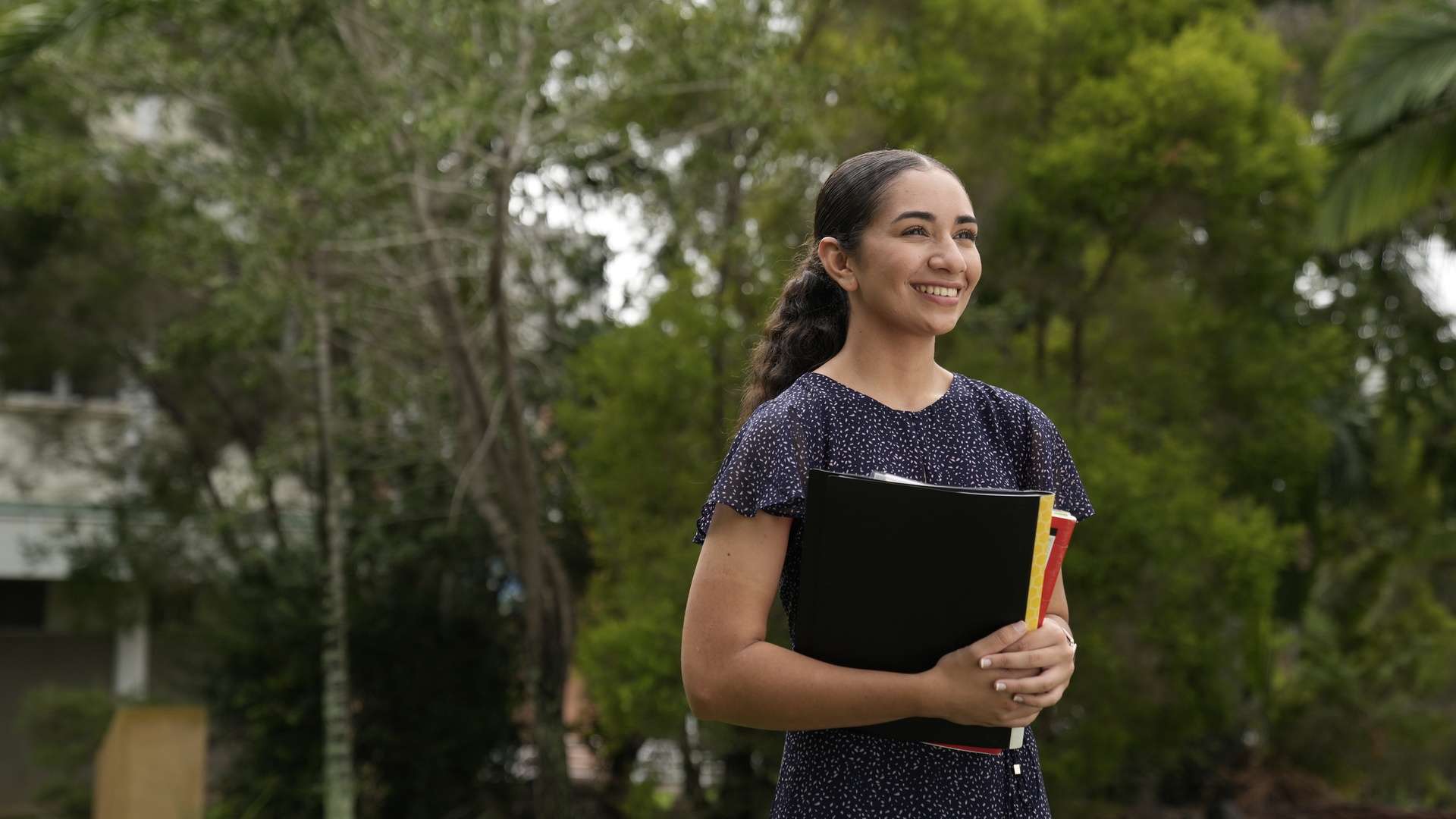 A First Nations student standing and holding textbooks outside on campus