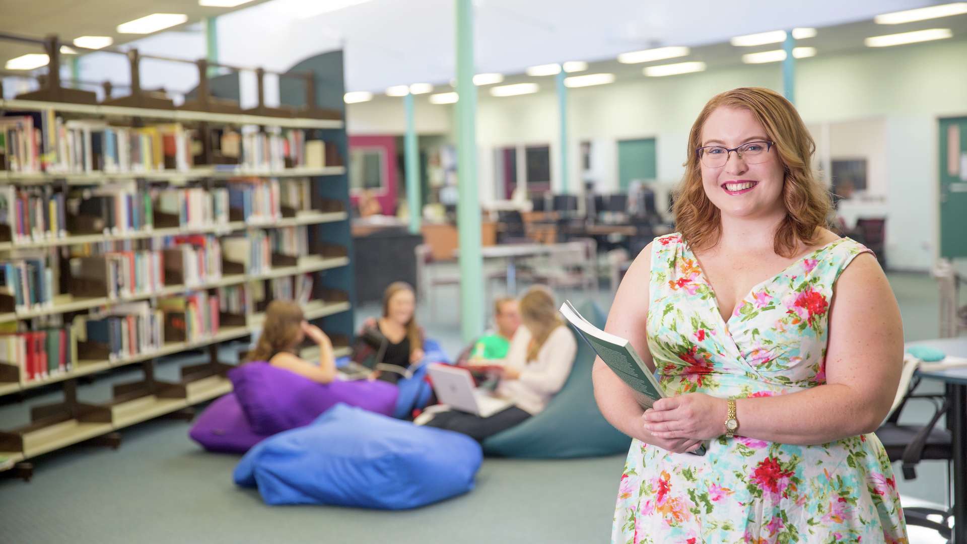 A happy student standing in a CQU library holding a textbook while a study group sits on beanbags in the background