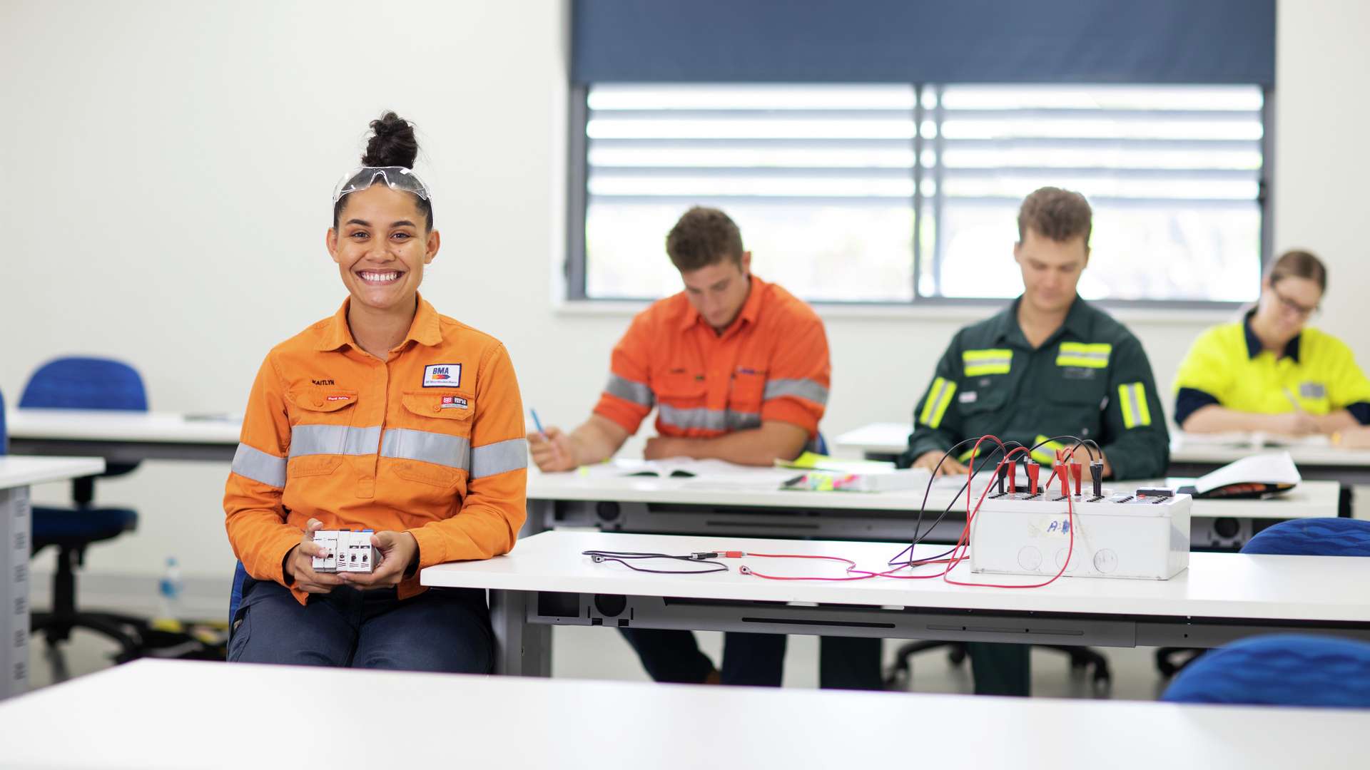 A trades student holding a circuit breaker in a classroom with other students studying in the background
