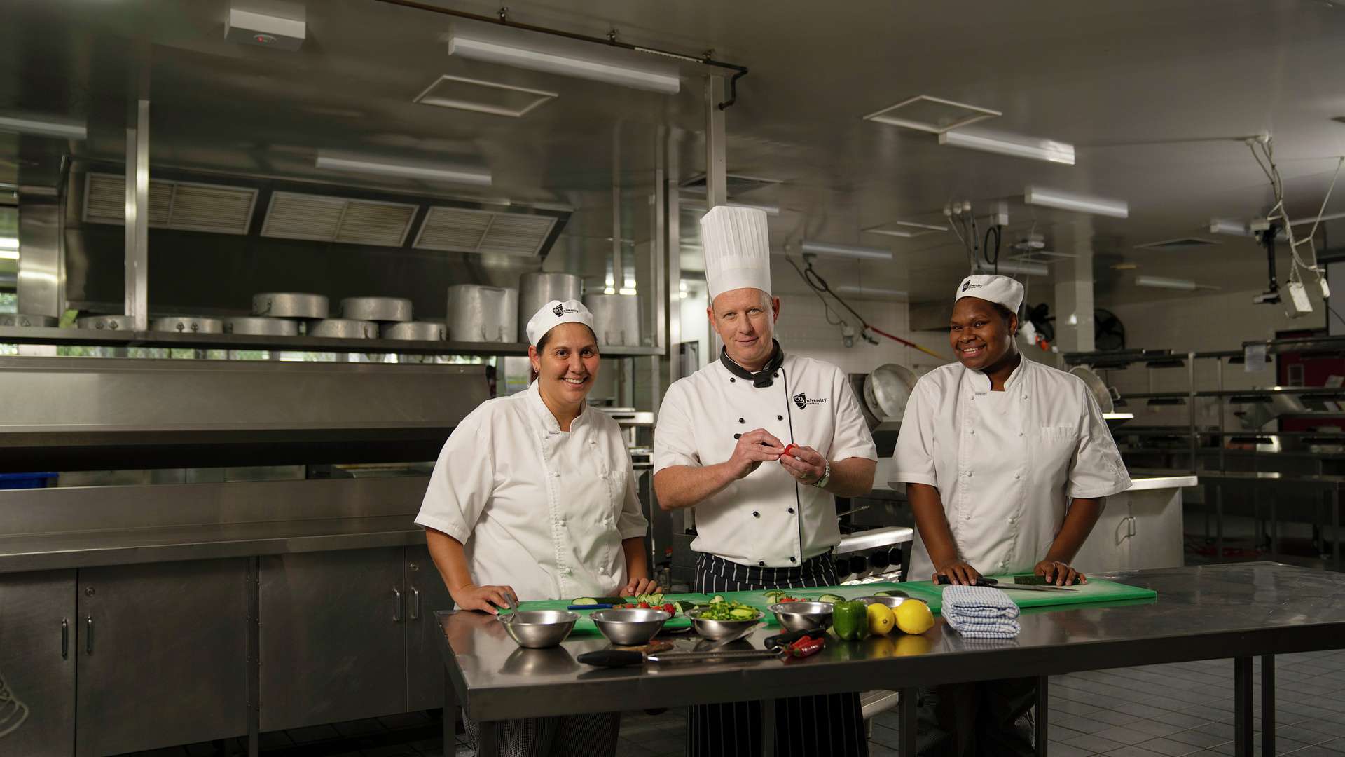 Two hospitality students and teacher in kitchen smiling at camera.