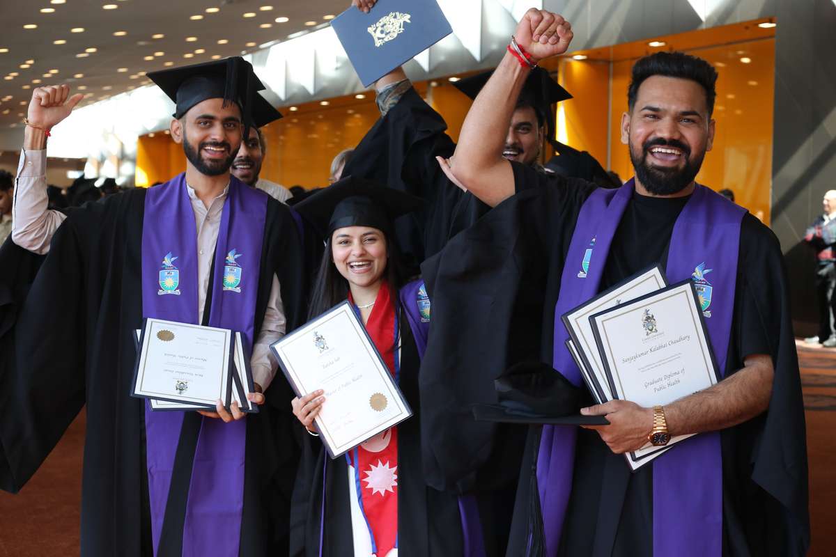 A group of graduates wear robes, hold testamurs, celebrate graduation.