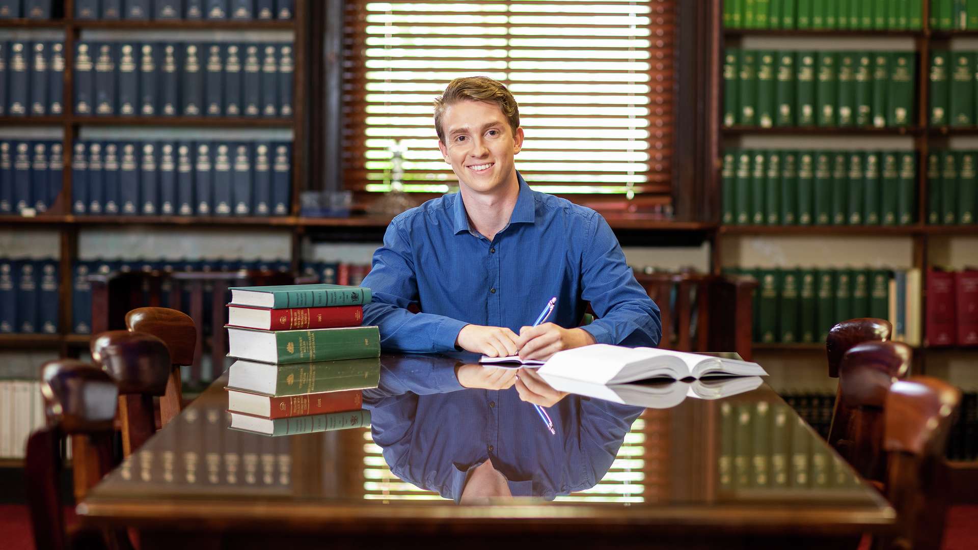 A Law student sits at a large mahogany desk in a library studying and smiling at the camera.