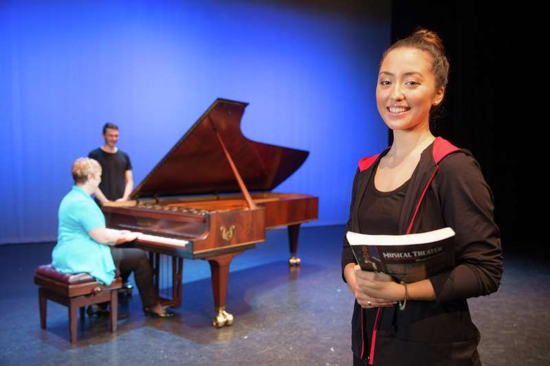 Musical theatre student holding a textbook on stage with lecturer playing piano in background