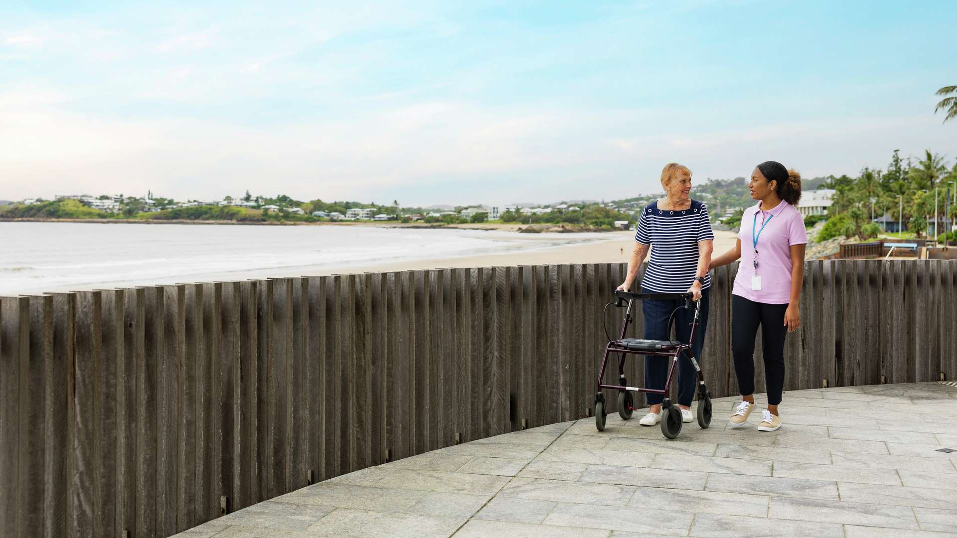 A CQU Community Services student takes a stroll along the esplanade at the beach with an elderly patient who requires assistance with walking