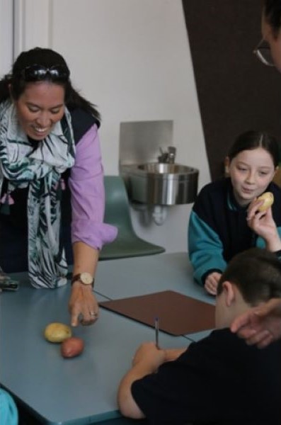 RACE Researcher pointing to potatoes on a desk as primary school children take notes around her