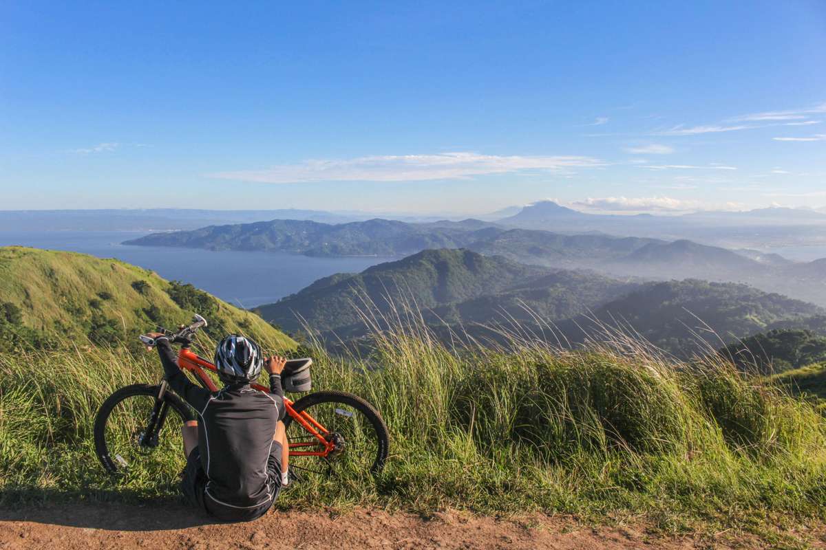 A person wearing a helmet sits beside a mountain bike on a grassy hilltop, looking out over rolling green mountains and a blue coastal landscape under a clear sky.