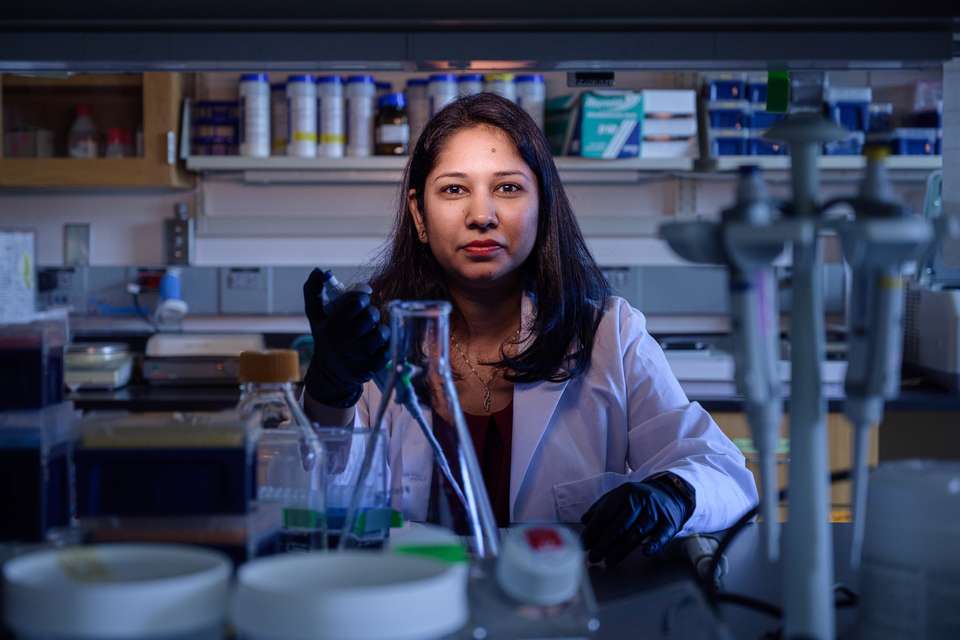 Dr Riti Sharan wears a lab coat and black gloves, holds lab equipment in a laboratory.