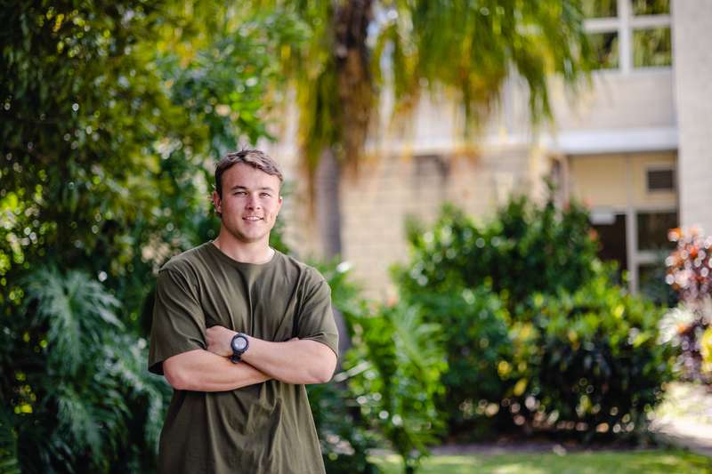 Student standing on campus with arms crossed smiling with gardens and building in background.