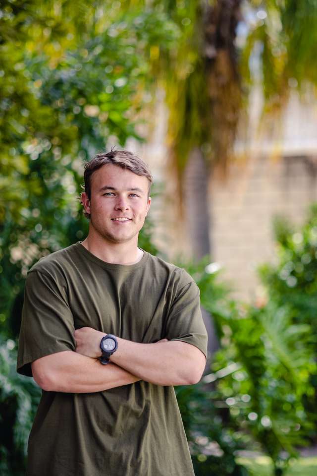 Student standing on campus with arms crossed smiling with gardens and building in background.