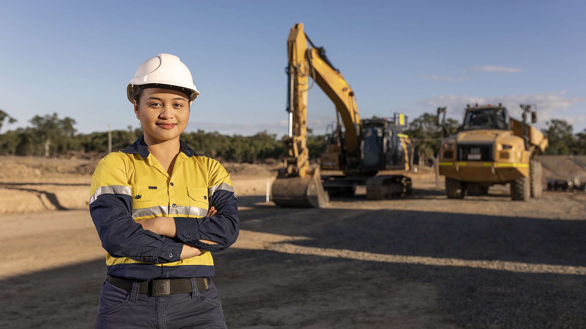 Engineering worker in hi vis and a hard hat stands in front of a worksite and machinery.