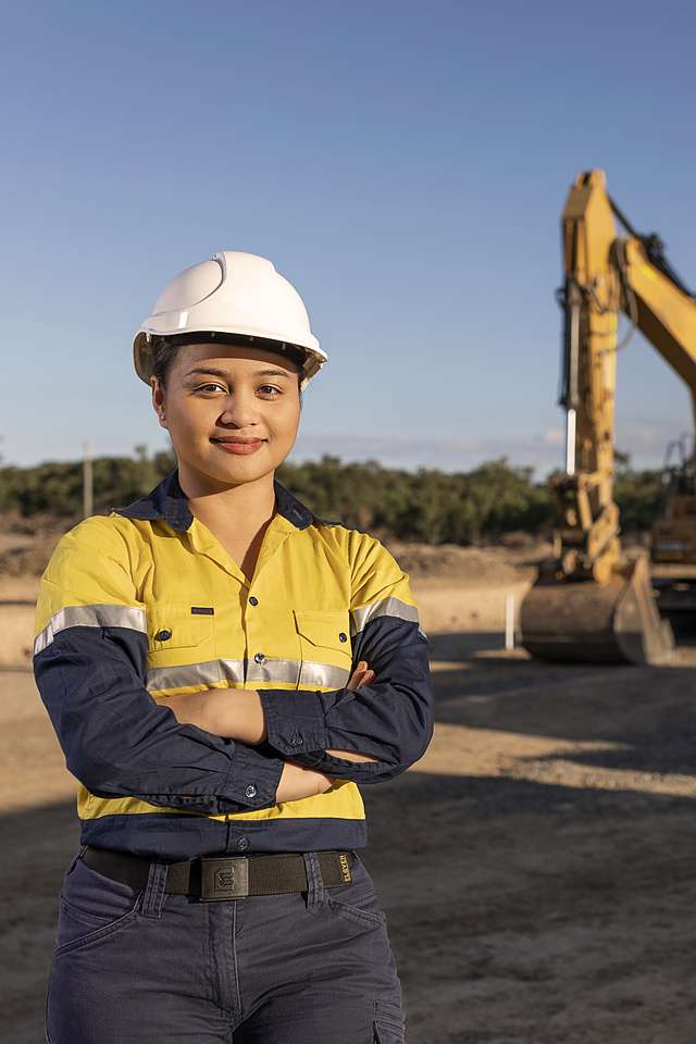 Engineering worker in hi vis and a hard hat stands in front of a worksite and machinery.