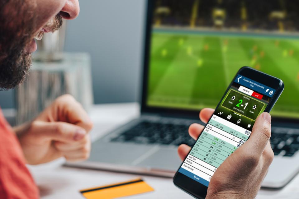 Person eagerly looking at their phone with a gambling app open as a soccer game plays on a laptop in the background