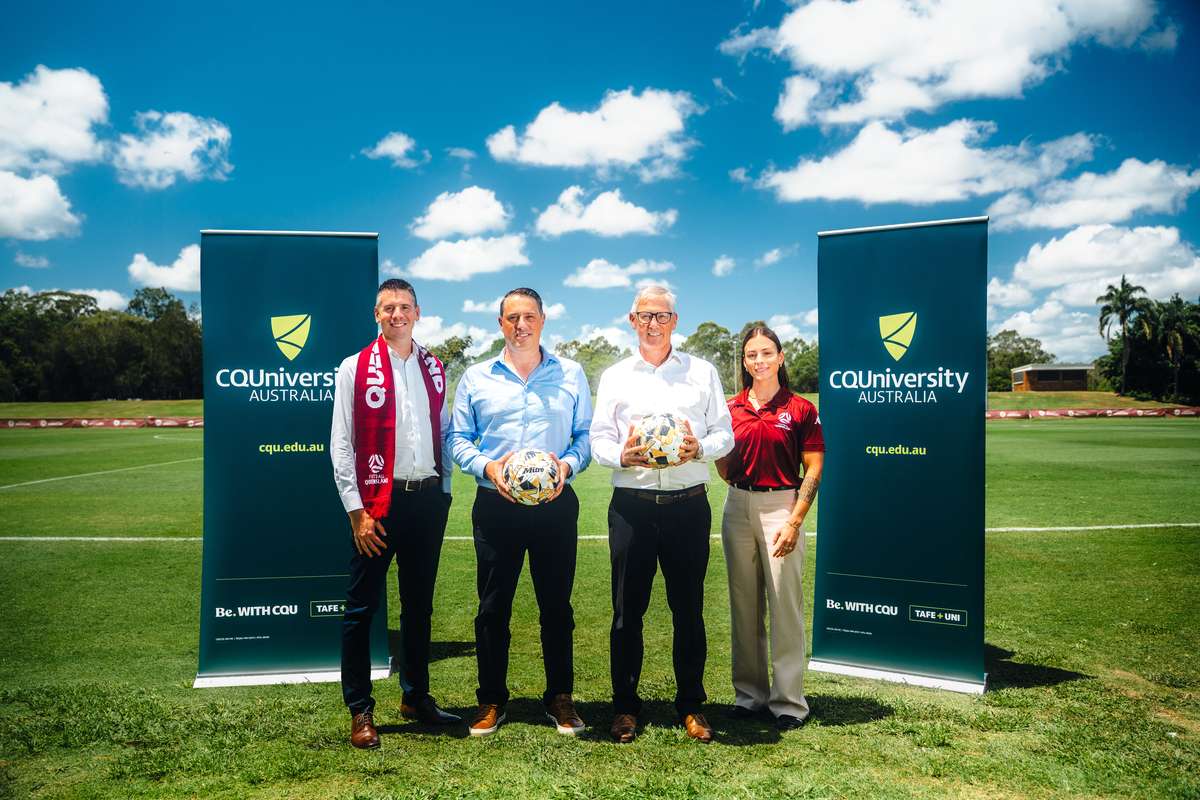 Four people stand on a football pitch, two men hold round footballs. The group is between two CQU banners.