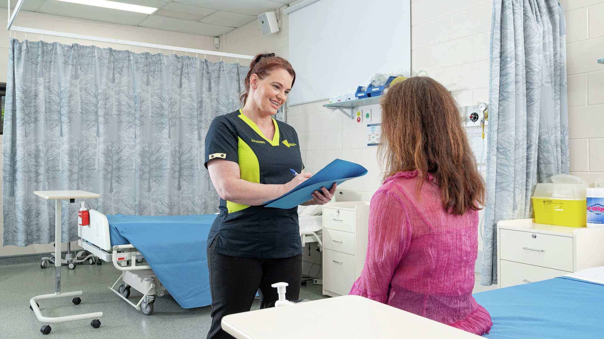 Nurse talks to patient holding a clipboard in a hospital ward.