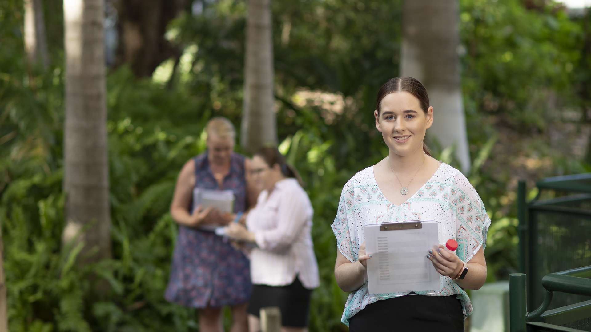 Student in the foreground with a clipboard standing in nature, with two other students in the distance.