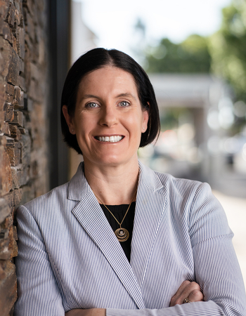 Headshot of Barbie Keller smiling in a pin striped jacket leaning against a brick wall