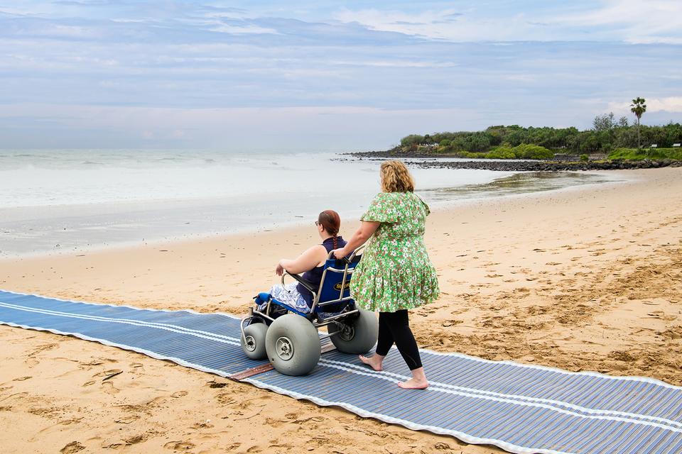 One person pushing another person towards the ocean in a wheelchair that has been designed for the sand