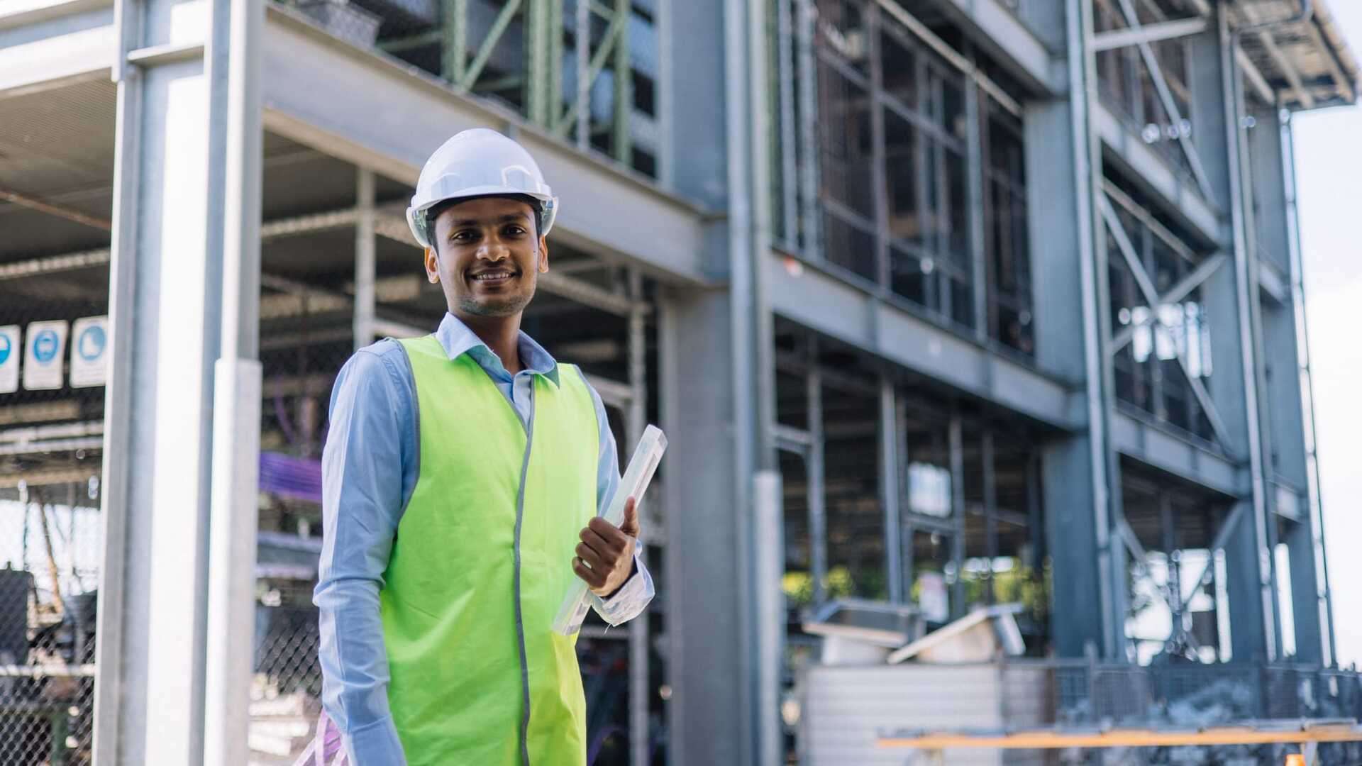 A Building Surveying student wearing PPE is holding paperwork in front of a construction site